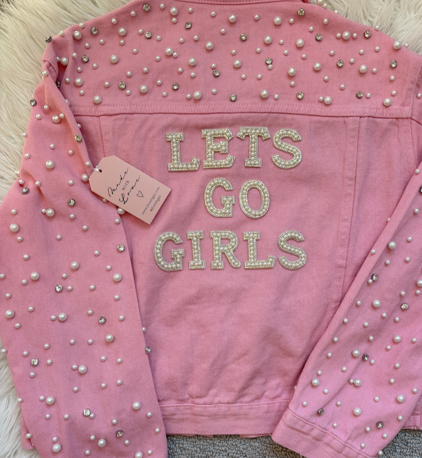 Pink jacket with pearl embellishments and 'LET'S GO GIRLS' text on a fluffy surface.